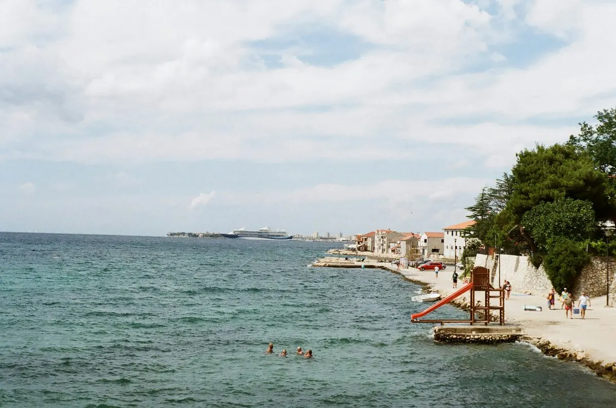 A seafront promenade at Bibinje with swimmers in the water and a cruise ship passing in the distance